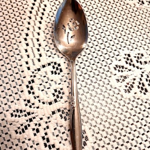 May include: A silver serving spoon with a floral design cut into the bowl. The spoon has a long, slender handle and is resting on a white lace tablecloth. The floral design is a stylized flower.