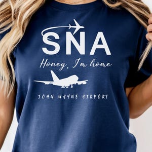 May include: Navy blue t-shirt with white text and graphics. The shirt features the letters "SNA" above the phrase "Honey, I'm home" and a graphic of an airplane. Below the text is "JOHN WAYNE AIRPORT".