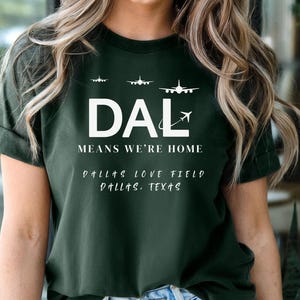 May include: Dark green t-shirt with white text and airplane graphics. The shirt reads "DAL MEANS WE'RE HOME" with "Dallas Love Field Dallas, Texas" below. The shirt has rolled-up sleeves and is worn by a person.