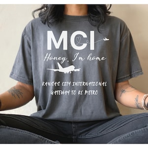 May include: A gray t-shirt with the text "MCI Honey, I'm home" and an airplane graphic. Below the text is "Kansas City International Gateway to KC Metro". The shirt is worn by a person sitting down.