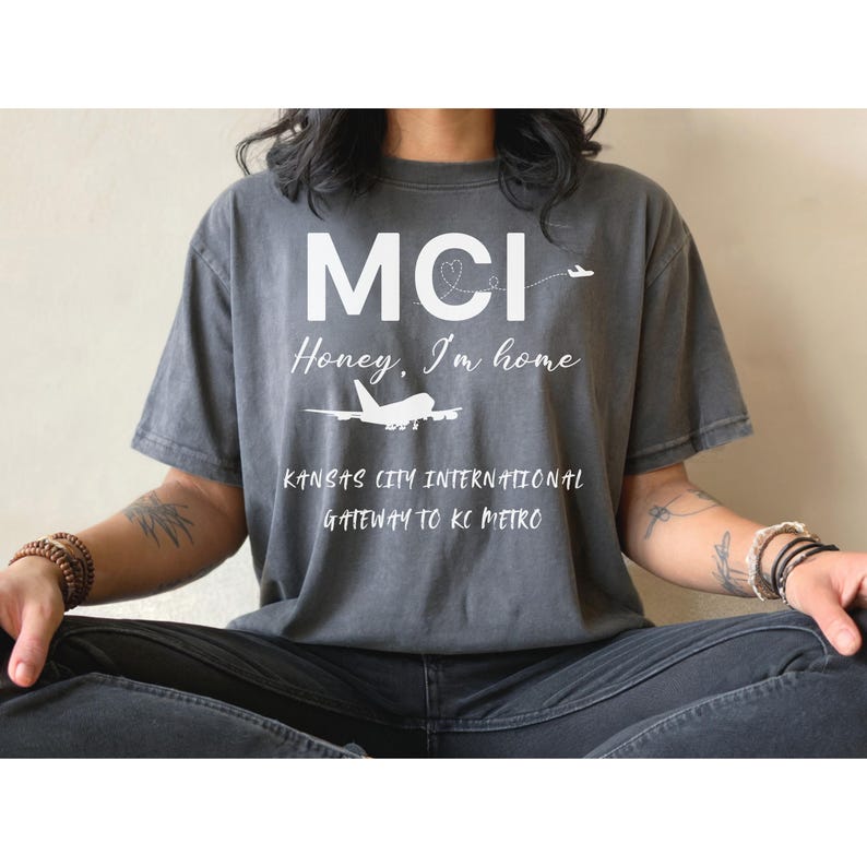 May include: A gray t-shirt with the text "MCI Honey, I'm home" and an airplane graphic. Below the text is "Kansas City International Gateway to KC Metro". The shirt is worn by a person sitting down.