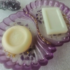 May include: A purple and white resin soap dish with two compartments. One compartment has a round white soap bar and the other compartment has a rectangular white soap bar.