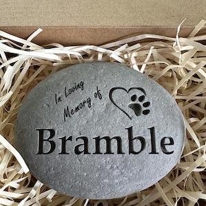 May include: A gray stone with the text "In Loving Memory of Bramble" and a paw print heart engraved on it. The stone is in a box with shredded paper.