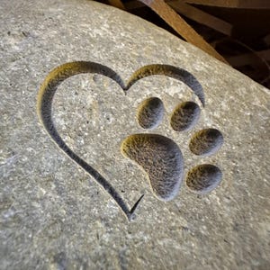 May include: A gray stone heart with a paw print carved inside. The heart is a symbol of love and the paw print represents a pet.