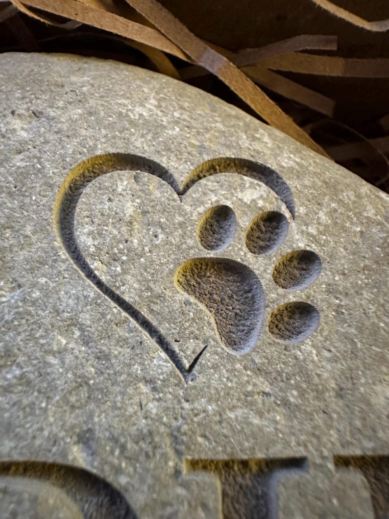 May include: A gray stone heart with a paw print carved inside. The heart is a symbol of love and the paw print represents a pet.