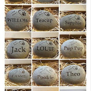 May include: A collection of 15 gray, round, engraved stones with paw prints and hearts. Each stone has a different name engraved on it, such as Bailey, Blackie, Gus, Willow, Teacup, Sapphie, Jack, Louie, Pup Pup, Cassie, Pookie, Theo, Bobby, Webber, and Bella Blot.