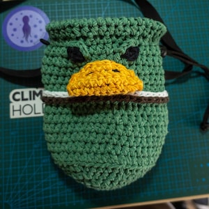 Puede incluir: Bolsa de magnesio de escalada de crochet verde con forma de pato, con un pico amarillo y ojos negros.