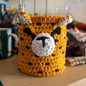 Peut inclure: Un récipient de rangement fait à la main, en crochet, en forme de guépard. Le récipient est principalement jaune avec des taches noires, des accents blancs et une sangle noire. Le visage du guépard est blanc avec des détails noirs.