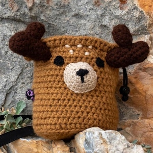 Peut inclure: Un sac à magnésie marron crocheté en forme de cerf, avec des bois marron foncé et un visage beige clair. Le cerf a des yeux noirs et un nez noir. Le sac a une fermeture à cordon et une sangle noire.