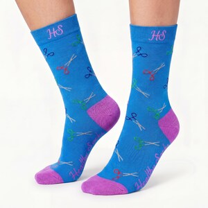 May include: A pair of bright blue crew socks featuring a repeating pattern of small scissors in red, green, and dark blue. The socks have purple toes and heels, with the letters HS embroidered in pink at the top and the text Healthy Socks in pink.