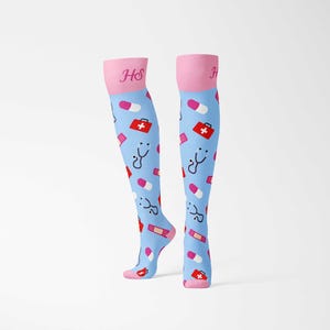 Könnte beinhalten: Kniestrümpfe mit hellblauem Hintergrund und medizinischen Symbolen. Die Socken haben rosa Akzente an der Spitze und den Zehen, mit den Initialen "HS" in Rosa. Das Muster umfasst rote Erste-Hilfe-Kästen, Stethoskope, Pillen und Verbände. Maße in cm.