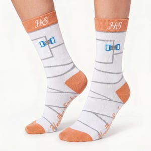 Può includere: Calze bianche con bordo arancione e le lettere HS ricamate in bianco. Il motivo presenta linee grigie che ricordano delle bende, piccoli accenti rettangolari blu e la scritta Healthy Socks in corsivo arancione sul lato della calza.