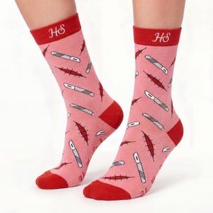 May include: A pair of pink crew socks featuring a repeating pattern of silver scalpels and red stitched surgical incisions. The socks have a solid red cuff with the white embroidered letters HS, and a solid red heel and toe section.