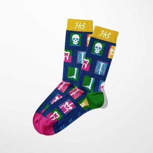Op de afbeelding: Een paar novelty sokken met een donkerblauwe achtergrond en een patroon van medische afbeeldingen, waaronder schedels, tanden en röntgenfoto's. De teen is roze, de hiel is groen en de boord is geel met de initialen "HS".