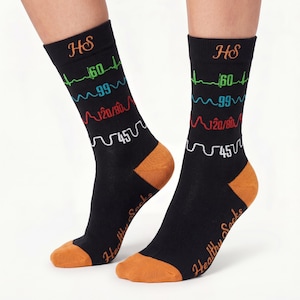 May include: Black crew socks featuring orange heels and toes with the text Healthy Socks. The design includes medical graphics of heart rate, oxygen saturation, blood pressure, and respiratory rate with the letters HS at the top of each sock.