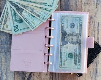 May include: A pink personal planner with a clear plastic pocket on the right side. A twenty dollar bill is inserted into the pocket. A stack of cash is on the left side of the planner.
