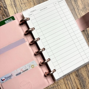 May include: A pink leather planner with a clear plastic insert for a budget tracker. The insert has columns for date, category, description, +/- and balance. The planner has six rose gold rings and a pocket on the front cover.