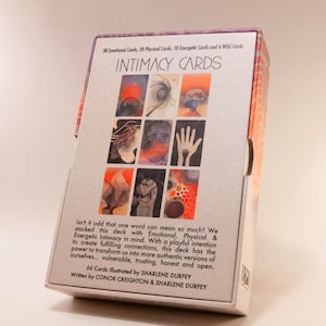 Może przedstawiać: Pudełko z kartami intymności z białym tłem i kolorową abstrakcyjną grafiką z przodu. Pudełko zawiera napis "INTIMACY CARDS" czarnymi literami. Na tylnej stronie pudełka znajduje się tekst: "Isn't it odd that one word can mean so much? We stacked this deck with Emotional, Physical & Energetic Intimacy in mind. With a playful intention to create fulfilling connections, this deck has the power to transform us into more authentic versions of ourselves... vulnerable, trusting, honest and open. 66 Cards Illustrated by SHARLENE DURFEY Written by CONOR CREIGHTON & SHARLENE DURFEY"