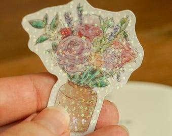 May include: A die-cut sticker featuring a watercolour illustration of a bouquet of pink, purple, and green flowers arranged in a tan ceramic vase. The sticker has a holographic finish with small, reflective sparkles across the entire surface.