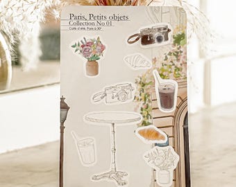 May include: A sticker sheet titled Paris, Petits objets Collection No 01 featuring illustrations of a brown camera, a bouquet of flowers in a vase, a croissant, an iced coffee, a round bistro table, and a street lamp on a light background.