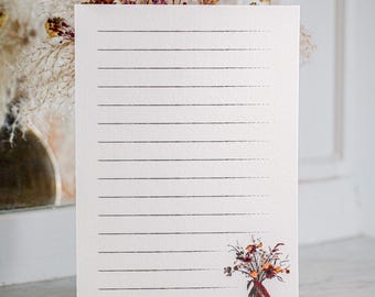May include: A rectangular sheet of white stationery paper featuring horizontal black lines for writing. The bottom right corner displays a small, detailed illustration of a colourful bouquet of dried flowers tied with a red ribbon.