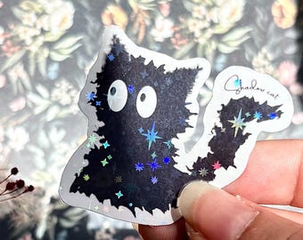 May include: A die-cut sticker of a fluffy black cat with large white eyes, featuring holographic star accents scattered across its body and tail. The text Shadow cat is printed in a cursive script on the tail, with a white border surrounding the design.