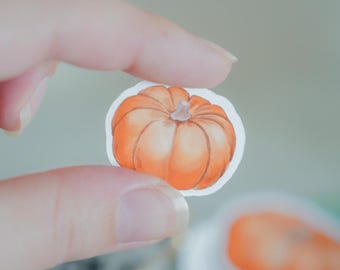 May include: A small, die-cut sticker featuring a watercolour-style illustration of a round, orange pumpkin with a light brown stem. The pumpkin has visible vertical ridges and a white border, presented as a single decorative adhesive item.