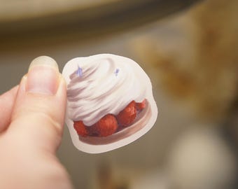 May include: A die-cut sticker featuring a detailed illustration of a dessert topped with a generous swirl of white whipped cream and bright red raspberries, accented with small blue sparkles on the cream surface.