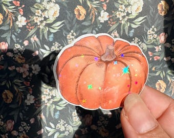 May include: A die-cut sticker of an orange pumpkin with a brown stem, featuring a holographic finish with small star-shaped accents scattered across the surface. The pumpkin has visible line shading and a rounded shape with distinct vertical ridges.