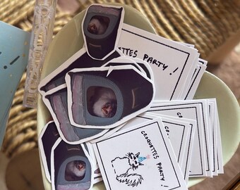 May include: A collection of stickers featuring a cat inside a carrier and a square sticker with a cat illustration and the text "CROQUETTES PARTY !" alongside a clear plastic bubble wand with a white rabbit-shaped topper in a light green bowl.