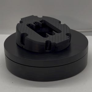 May include: A black, 3D-printed object with a circular base and a top section featuring a unique, slotted design. The top has a carbon fibre texture. The object is displayed on a black, circular platform.