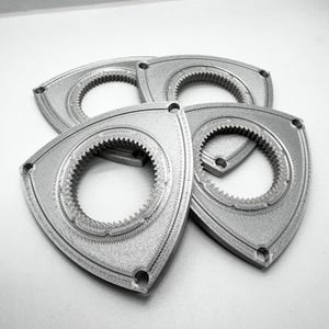May include: Four silver-coloured, triangular mechanical parts with a circular gear design in the centre. Each part has three holes and a textured surface. The parts are arranged in a slightly overlapping pattern.