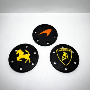 May include: Three black circular emblems with automotive logos. One has an orange arrow, another a yellow horse, and the third a yellow bull with the word "Lamborghini". Each emblem has six evenly spaced holes.