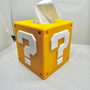 May include: A yellow tissue box featuring white question marks, designed to look like a video game block. White tissues protrude from the top. The box is square with rounded corners and small circular details.