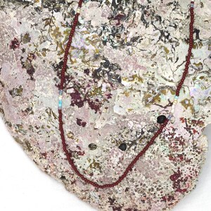 Puede incluir: Un delicado collar con pequeñas cuentas de color burdeos. El collar tiene un diseño sencillo con cuentas de acento blancas y azul claro. Se muestra sobre una superficie texturizada y multicolor. Una joya sencilla y elegante.
