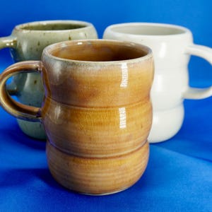 May include: Three ceramic mugs with rounded, stacked-ring bodies and side handles. The front mug is a glossy, crackled amber-brown. Behind it are two mugs: one is a speckled, muted sage green, and the other is a smooth, solid off-white.