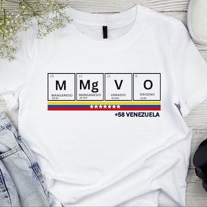 Puede incluir: Camiseta blanca con un diseño que presenta los elementos M, Mg, V y O, la bandera venezolana y el texto "+58 VENEZUELA". La camiseta tiene cuello redondo y mangas cortas.