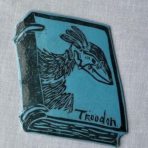 Może przedstawiać: Niebieska, prostokątna naklejka z czarną ilustracją ptasiego stworzenia w hełmie. Słowo "Troodon" jest wydrukowane na dole na czarno. Naklejka ma kształt książki.