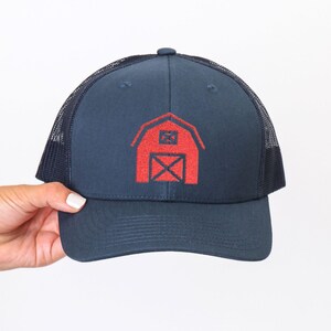 Può includere: Cappellino da camionista blu navy con grafica di un fienile rosso. Il cappello ha un retro in rete e una visiera curva. Il cappello è tenuto da una persona.