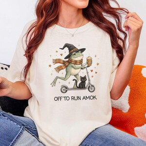 Puede incluir: Una camiseta color marfil con un gráfico de una rana verde con sombrero de bruja y bufanda de cuadros, montada en un patinete con un gato negro. La rana sostiene una taza de café, rodeada de pequeños corazones y estrellas naranjas, con el texto OFF TO RUN AMOK.