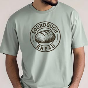 Op de afbeelding: Lichtgroen t-shirt met een cirkelvormig ontwerp. Het ontwerp bevat de woorden "SOURDOUGH BREAD" rond een tekening van een brood. Het t-shirt heeft korte mouwen en is effen van kleur.
