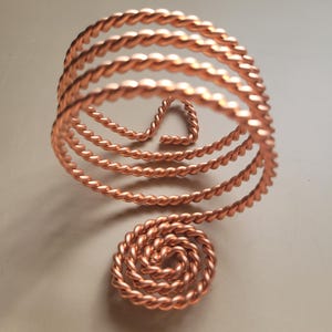 Op de afbeelding: Een koperkleurige, spiraalvormige armband met een gedraaid touwontwerp. De armband heeft meerdere concentrische cirkels, met een kleinere spiraal aan de basis. Het ontwerp is open en luchtig, met een unieke, handgemaakte uitstraling.
