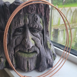 Op de afbeelding: Een koperen draadketting met een drievoudig strengontwerp. De ketting is opgerold en rust op een getextureerd, bruin, boomvormig beeld.