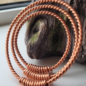 May include: Three copper wire bracelets, each with a twisted design, are stacked on top of each other. The bracelets are connected by a coiled copper wire.