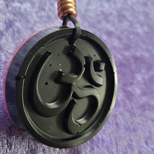 Puede incluir: Colgante negro con el símbolo Om en relieve, suspendido de un cordón negro. El colgante circular tiene un borde rosa y morado y un detalle de espiral de cobre en la parte superior.