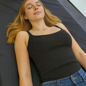May include: A black ribbed vest top with thin straps and blue denim jeans. The person is lying down with their eyes closed, and their blonde hair is spread out. The image is taken from a slightly elevated angle.