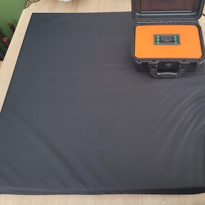 Può includere: Un grande tappeto rettangolare nero con una superficie strutturata. Una valigetta nera a guscio rigido aperta con interno arancione è appoggiata sul tappeto. La valigetta contiene un pannello di controllo con pulsanti verdi e un display digitale. Un cavo di alimentazione nero esce dalla valigetta.