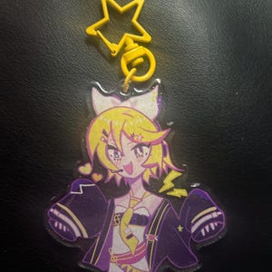 Może przedstawiać: Przezroczysty brelok akrylowy z postacią z kreskówki o żółtych włosach, fioletowych oczach i różowo-fioletowym stroju. Postać nosi żółty krawat i czarną kurtkę z gwiazdą na rękawie. Brelok ma żółty, gwiazdkowaty zawieszkę.