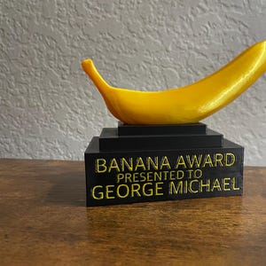 May include: A black award stand with a yellow banana sculpture on top. The stand has the text "BANANA AWARD PRESENTED TO GEORGE MICHAEL" in yellow lettering.