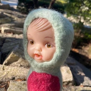 Pode incluir: Uma pequena boneca feita à mão com um traje de capuz verde e um círculo rosa na frente. A boneca tem um rosto de plástico com olhos castanhos e uma expressão sorridente.